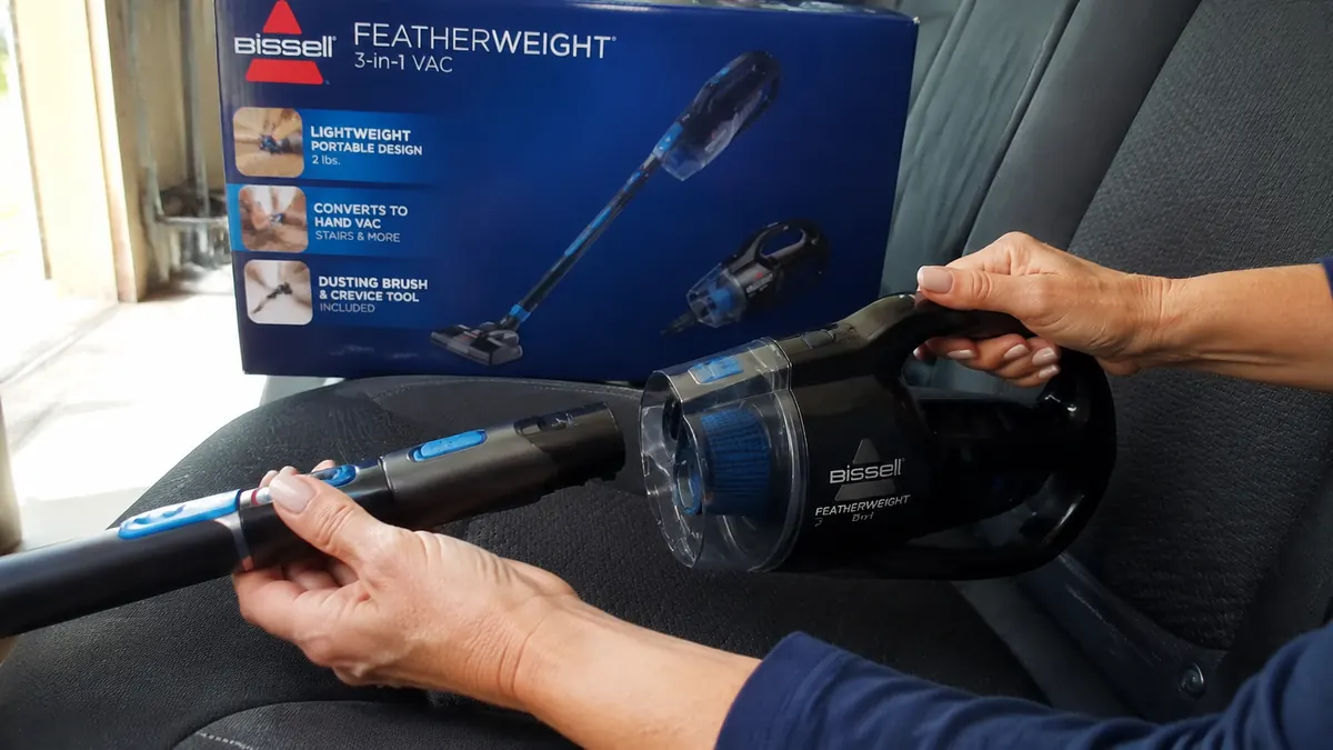 Hand detaching the Bissell Featherweight's upper wand to convert it into handheld mode above a car seat