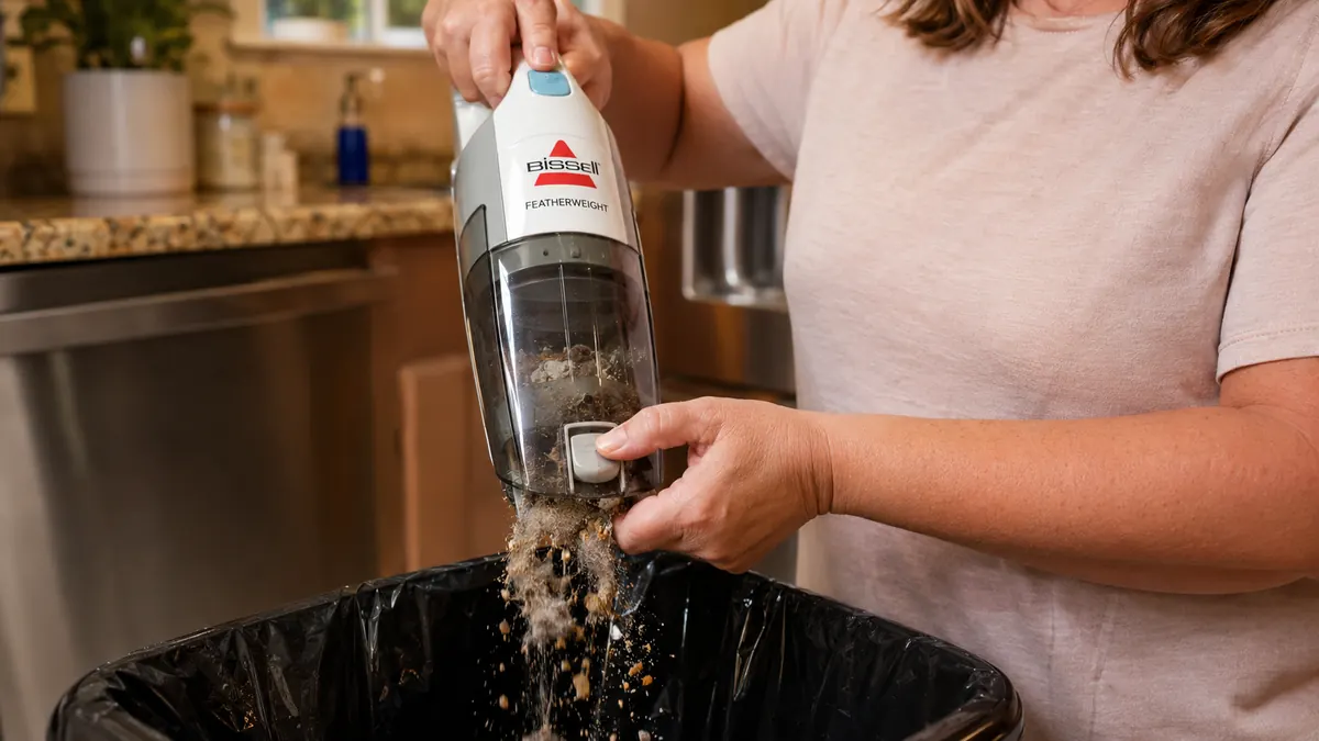 Close-up of the Bissell Featherweight's clear dust cup being emptied over a kitchen trash can, full of crumbs and pet hair