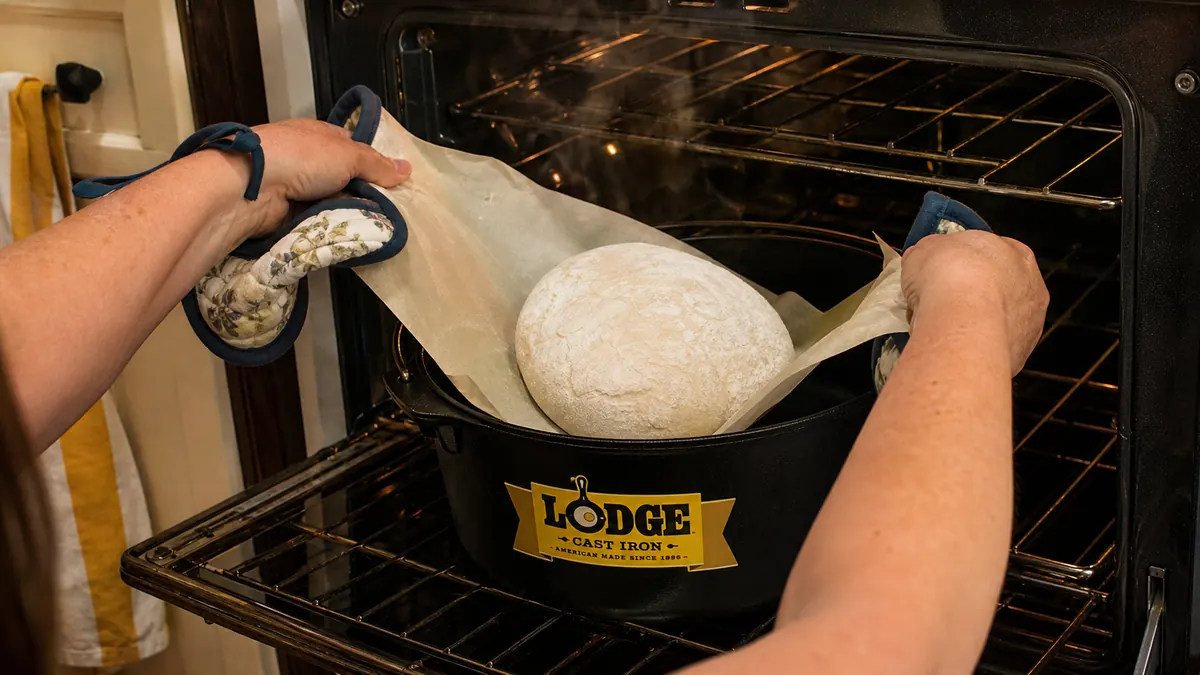 Hands lowering a parchment-lined dough ball into a preheated Lodge Dutch oven using the parchment as a sling
