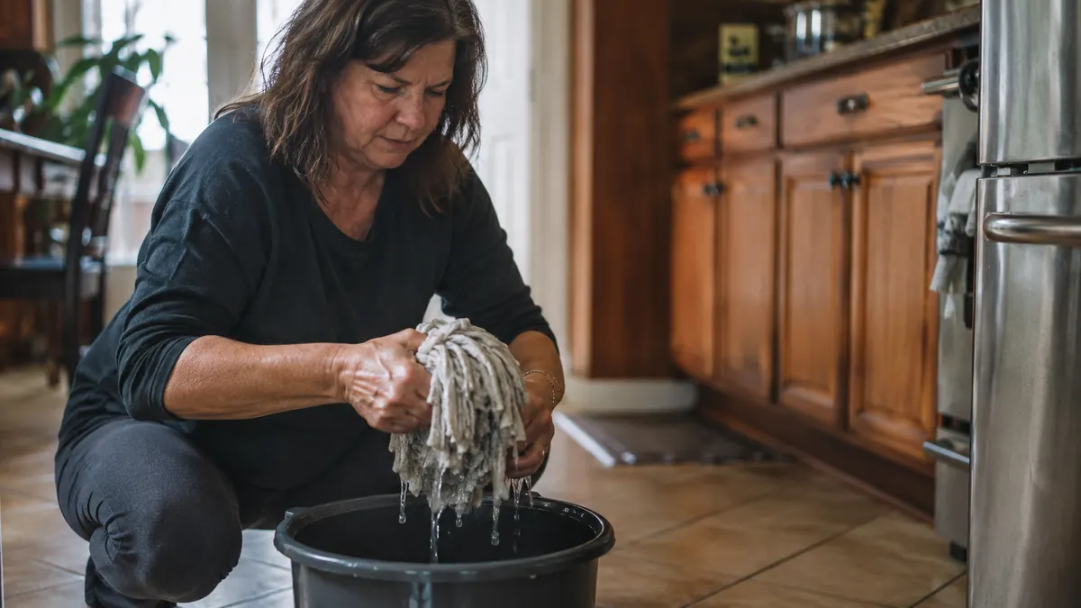 Damp mop finishing a tile floor after a stick vacuum pass, bucket and mop head visible near the kitchen island