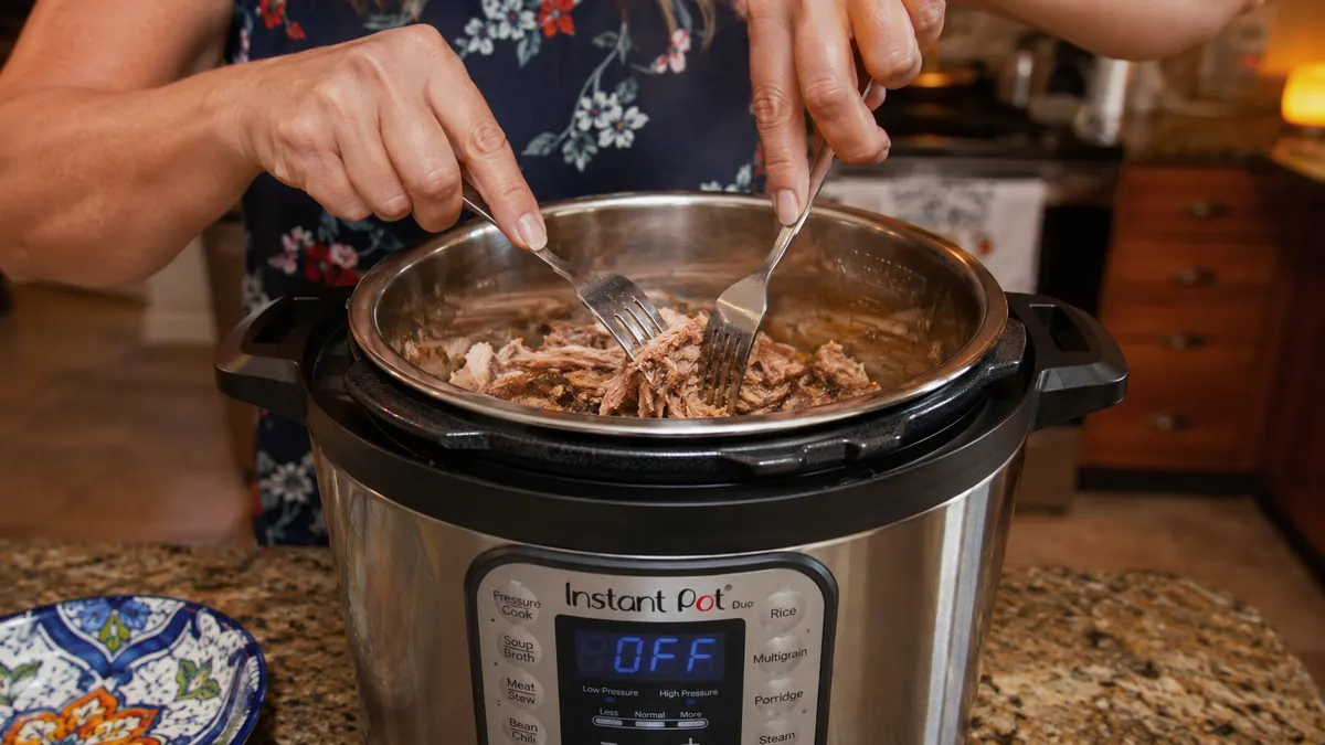 Two forks shredding cooked pork directly inside the stainless steel insert of an electric pressure cooker