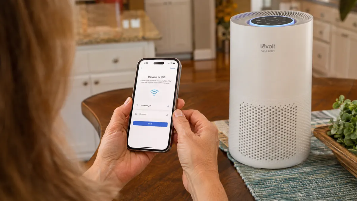 A woman's hands holding a phone showing the VeSync app setup screen next to the Levoit Vital 200S purifier